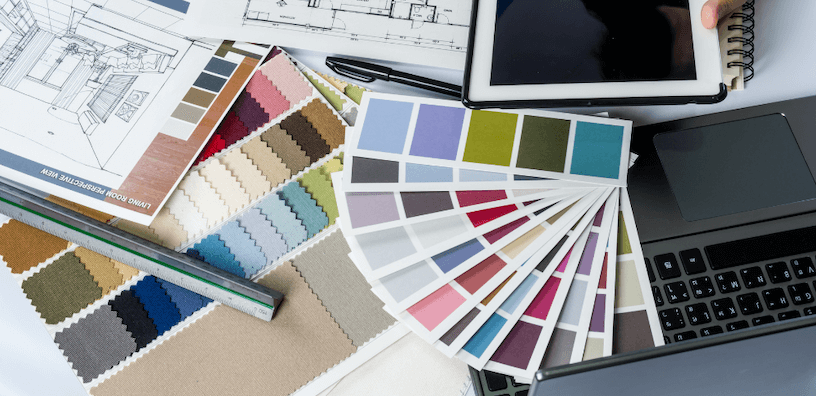 Une table recouverte d’échantillons de tissu, un ordinateur portable, une règle et un carnet de croquis de design d’intérieur.