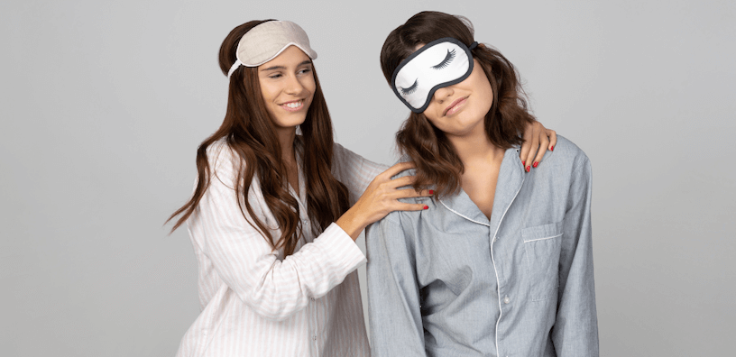 Deux femmes en pyjama, portant un masque de sommeil ; l’une semble gaie et alerte, tandis que l’autre a l’air endormie et semble avoir besoin d’être réconfortée, représentant des niveaux contrastés d’énergie matinale.