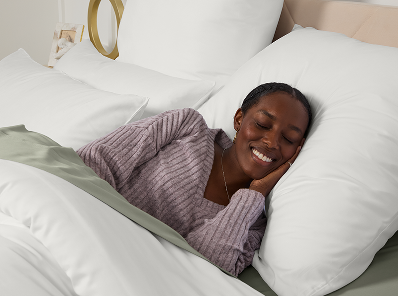Femme dormant paisiblement dans un lit avec des draps blancs et vert sauge, vêtue d’un pull lavande, souriante et détendue.