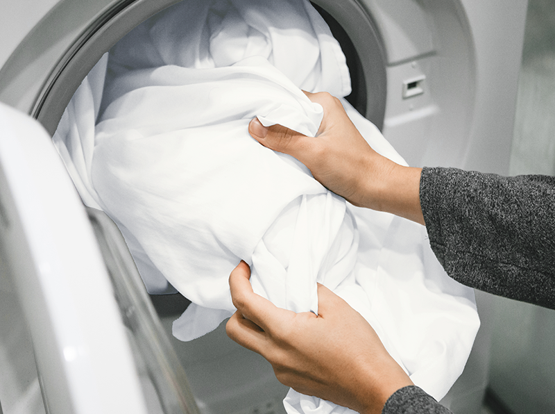 Mains chargeant des draps blancs doux dans une machine à laver, mettant en avant l’entretien facile.